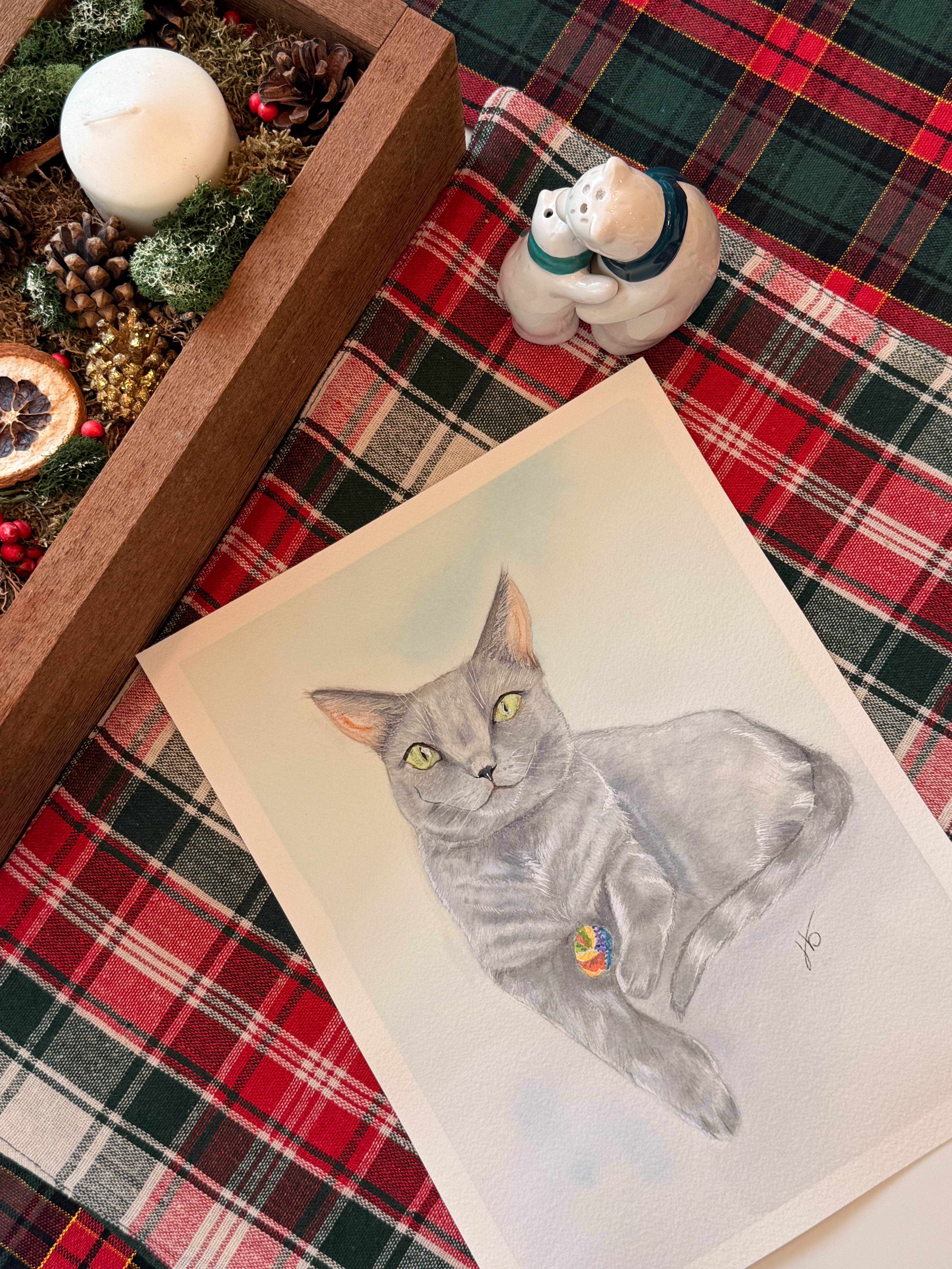 Colored pencil drawing of a gray cat lying with a small toy, photographed beside a Christmas centrepiece