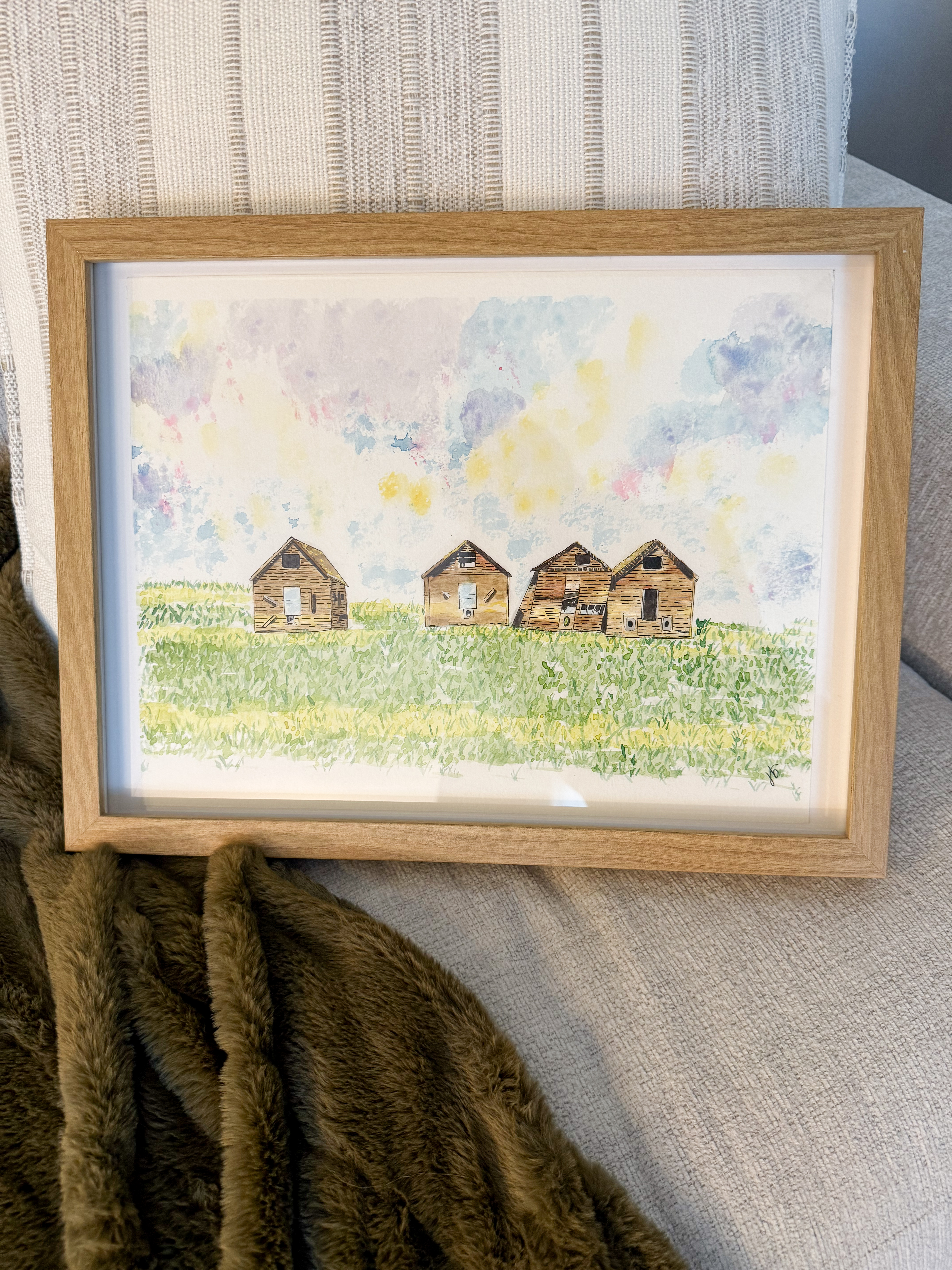 Framed watercolor of three weathered prairie homesteads in a green field