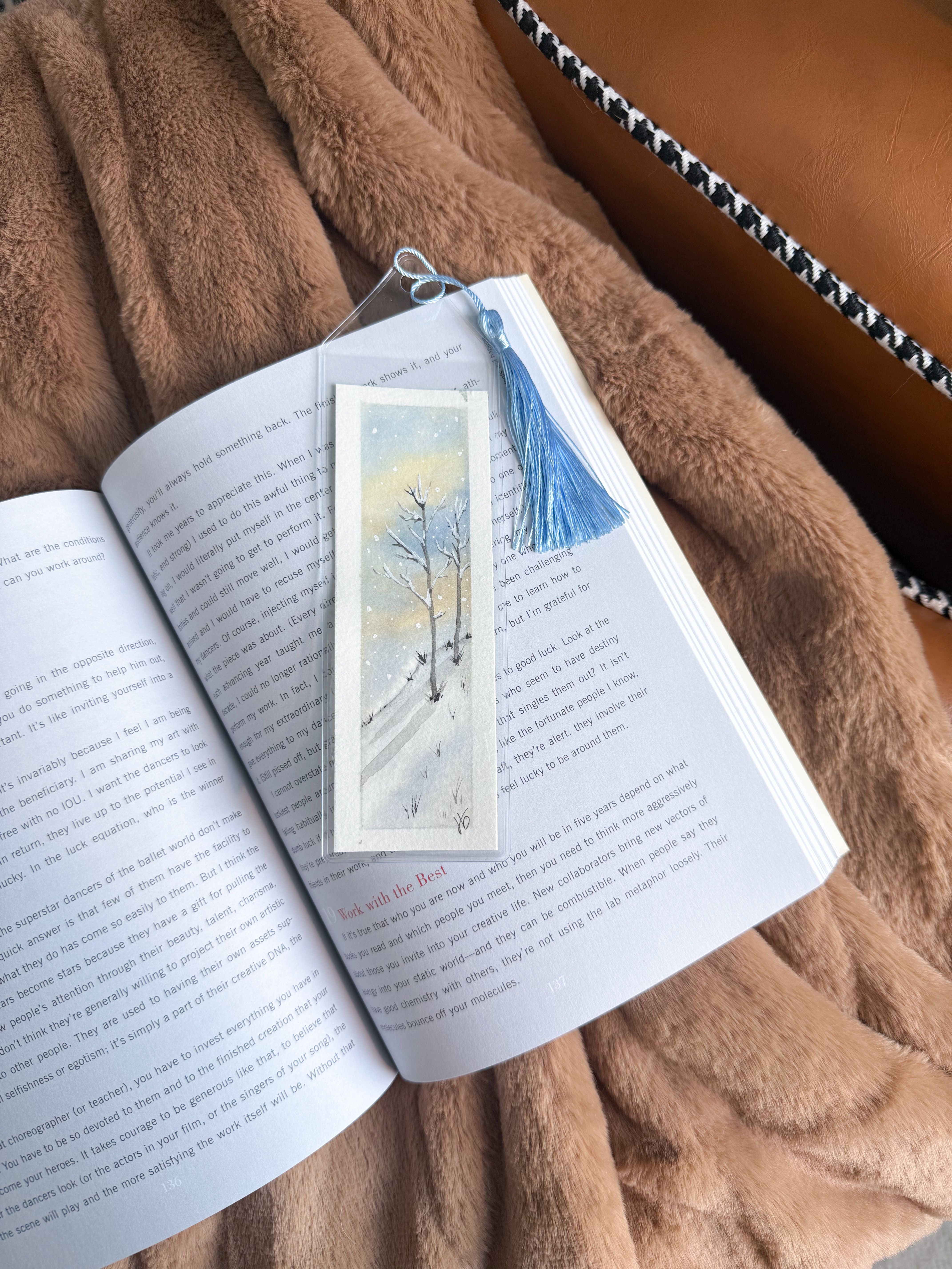 Watercolor bookmark of bare winter trees in snow, tucked into an open book with a blue tassel