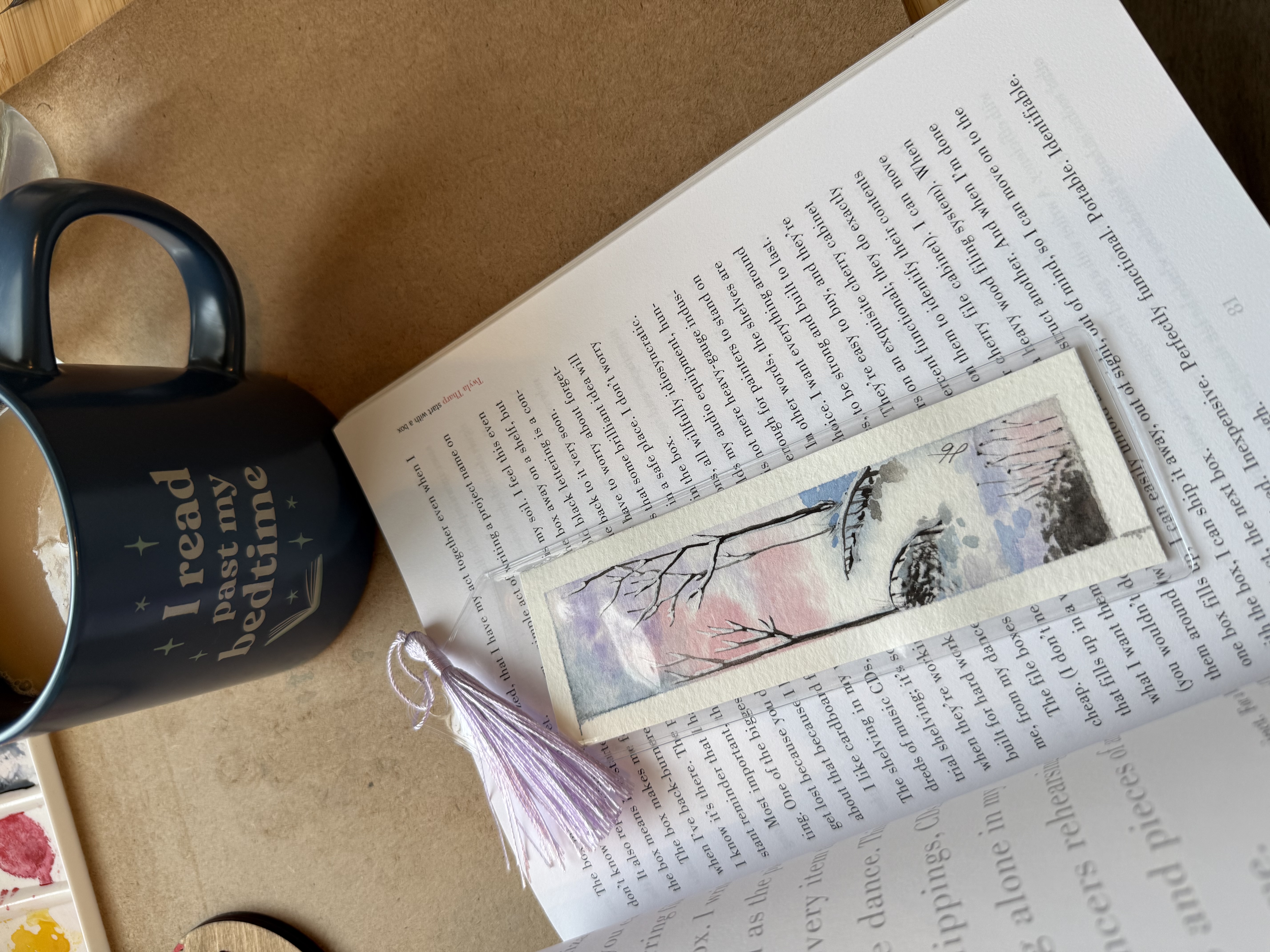 Abstract watercolor bookmark of a birch tree with a lavender tassel, set on an open novel