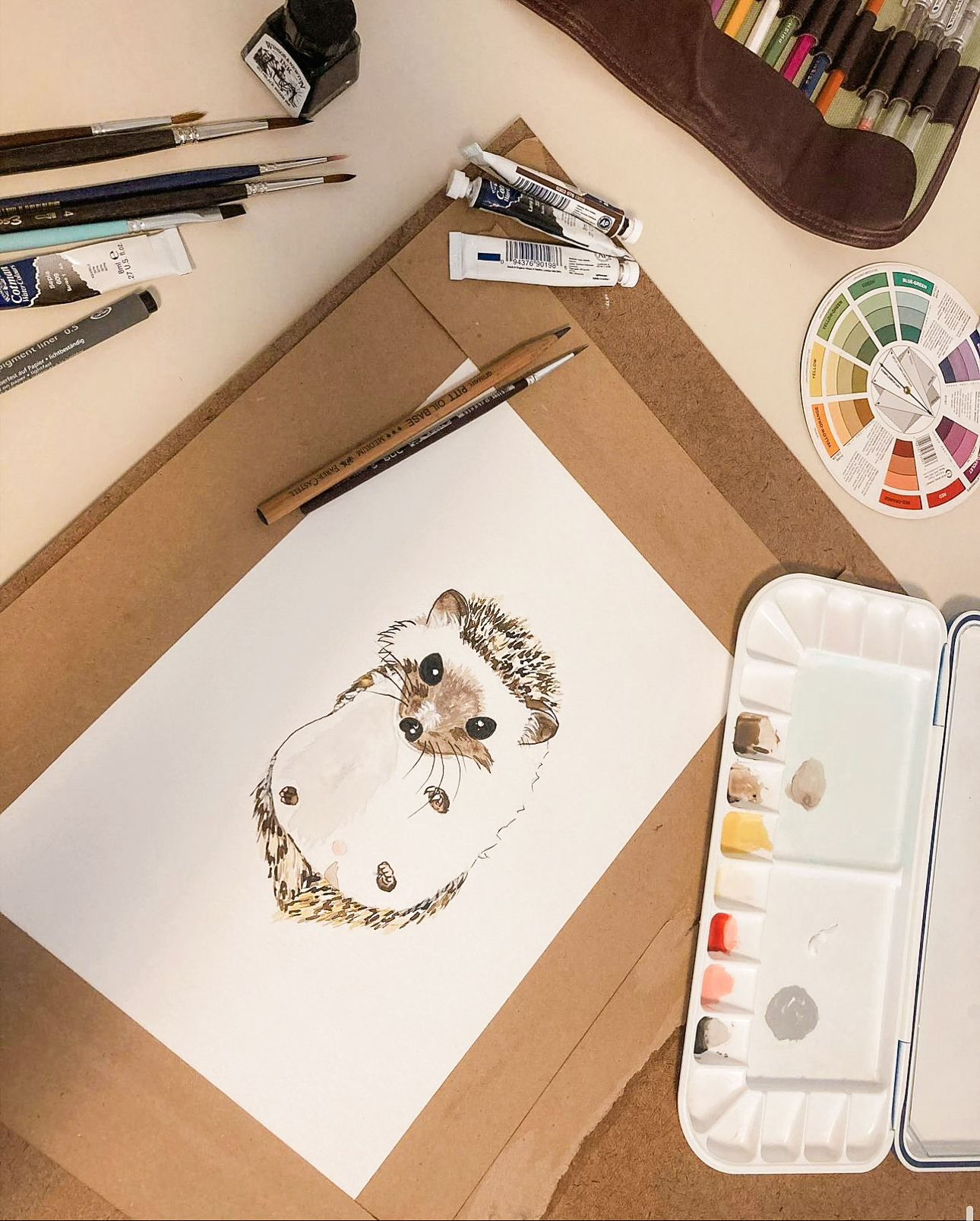 Watercolor of a hedgehog on a desk surrounded by paints, brushes and a colour wheel