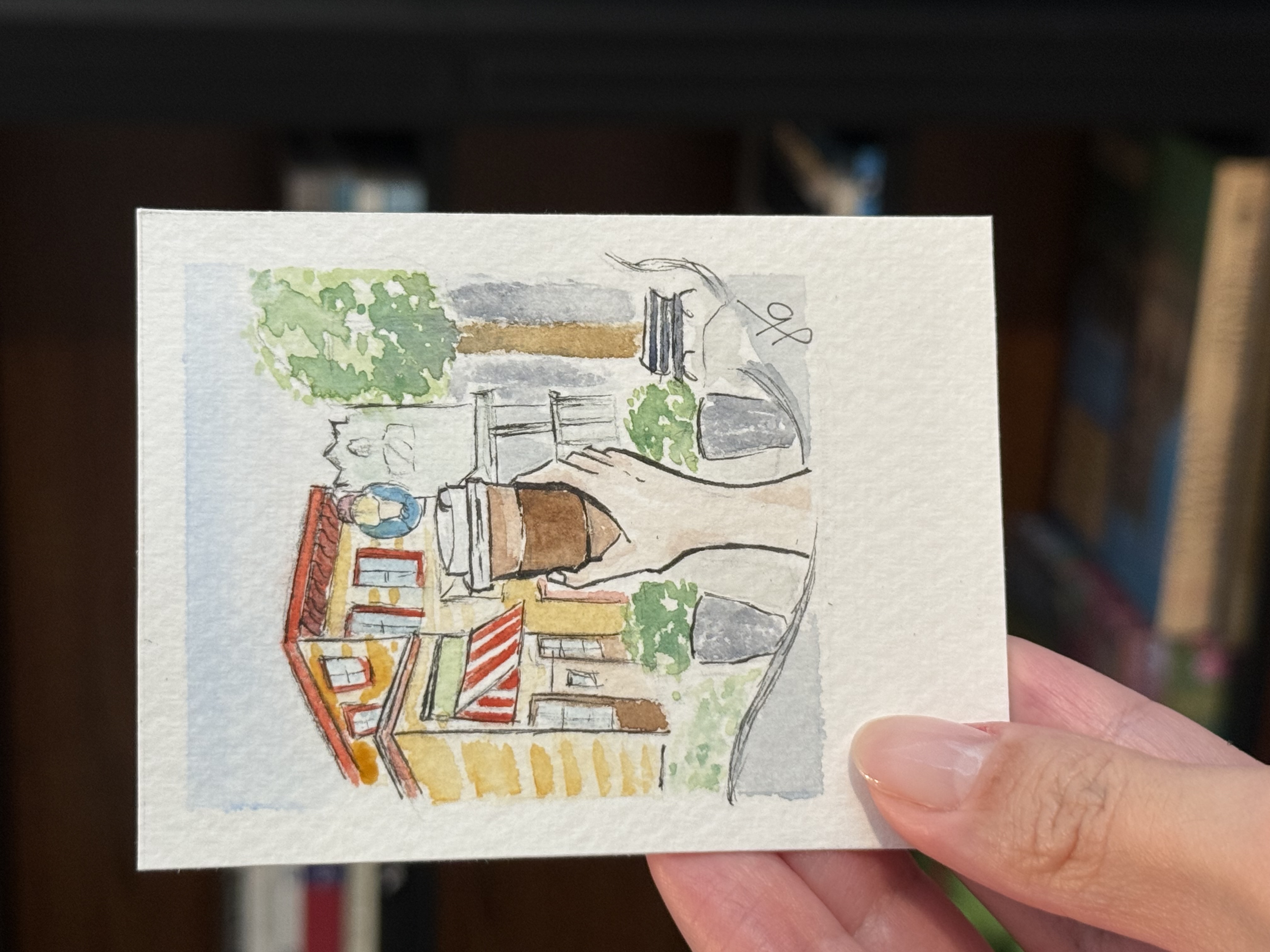 Tiny watercolor of a striped-awning sidewalk café held between fingertips
