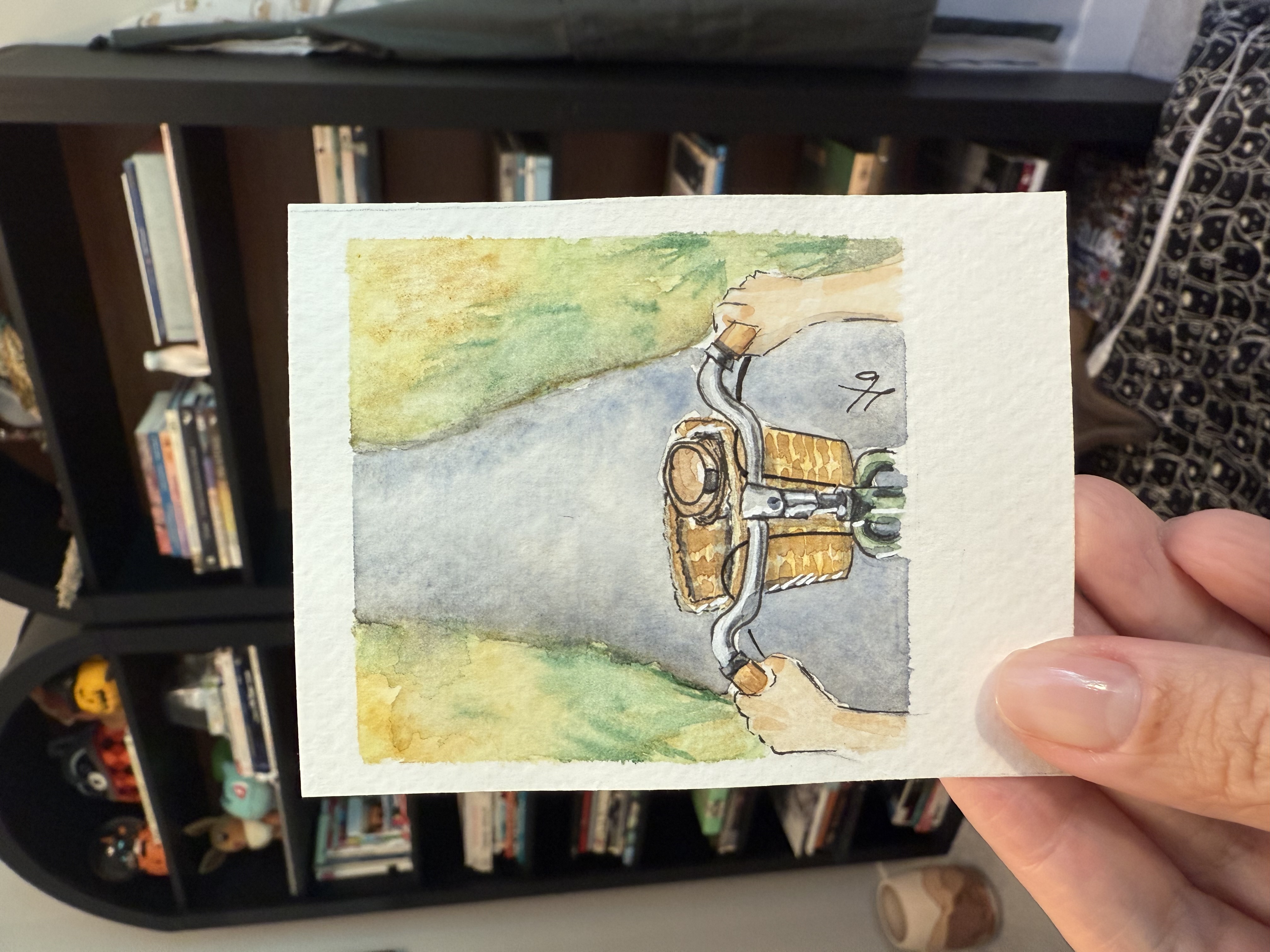 Tiny watercolor seen from a cyclist's POV with hands on bicycle handlebars and a basket up front
