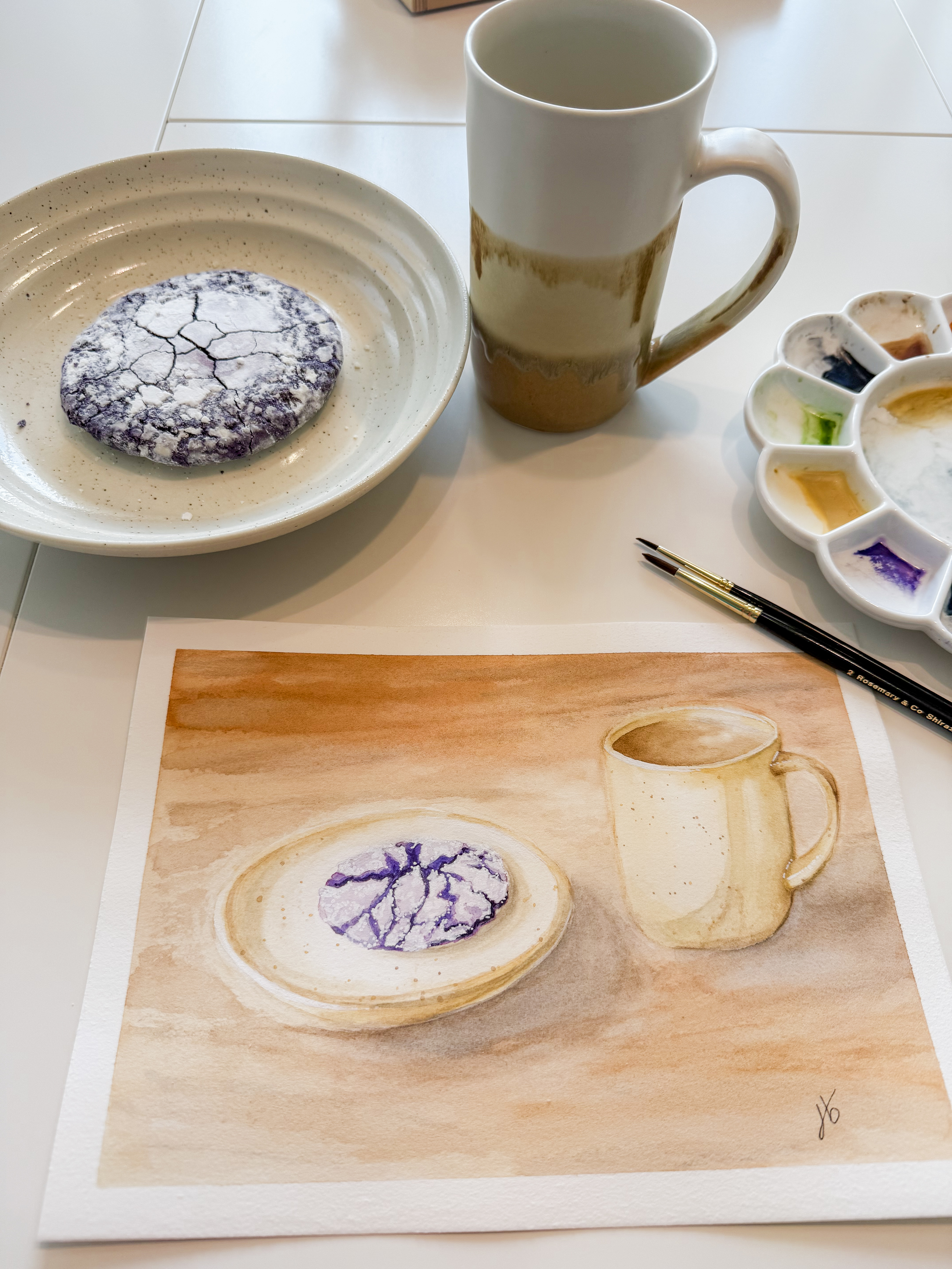Watercolor still life of a crinkle cookie and mug photographed beside the actual cookie, mug and palette